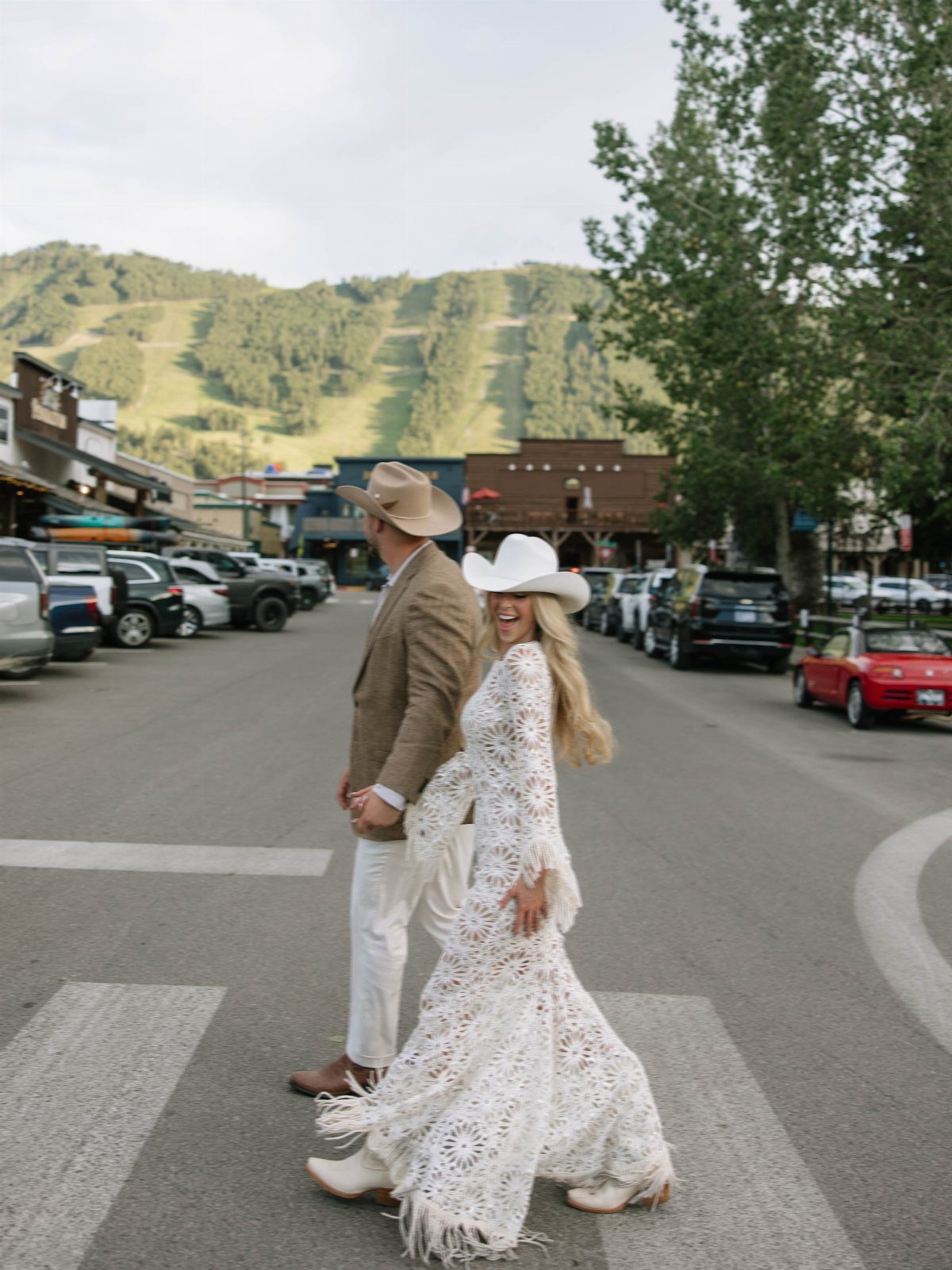 Bride and groom in western wedding attire walking across a mountain town street.