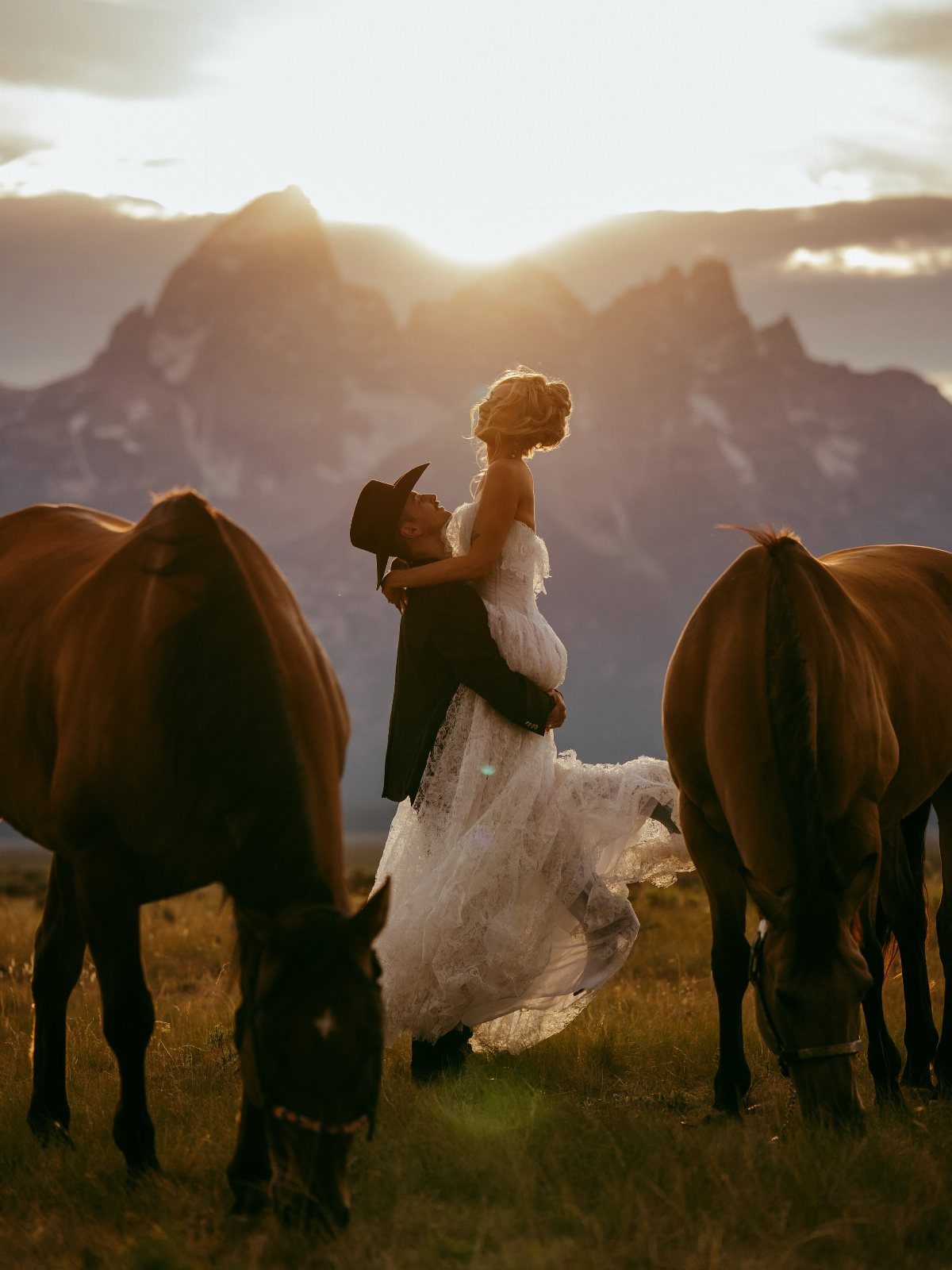 Bride and groom embracing in a field with horses and mountains at sunset.