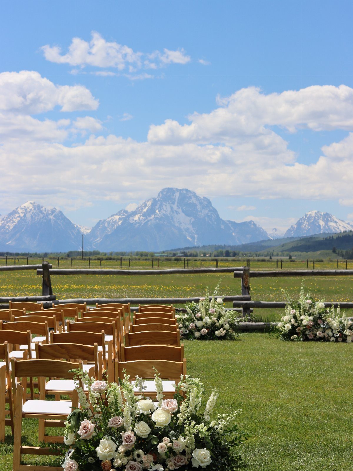 Wooden wedding chairs and floral arrangements facing snow-capped mountains in a grassy field.