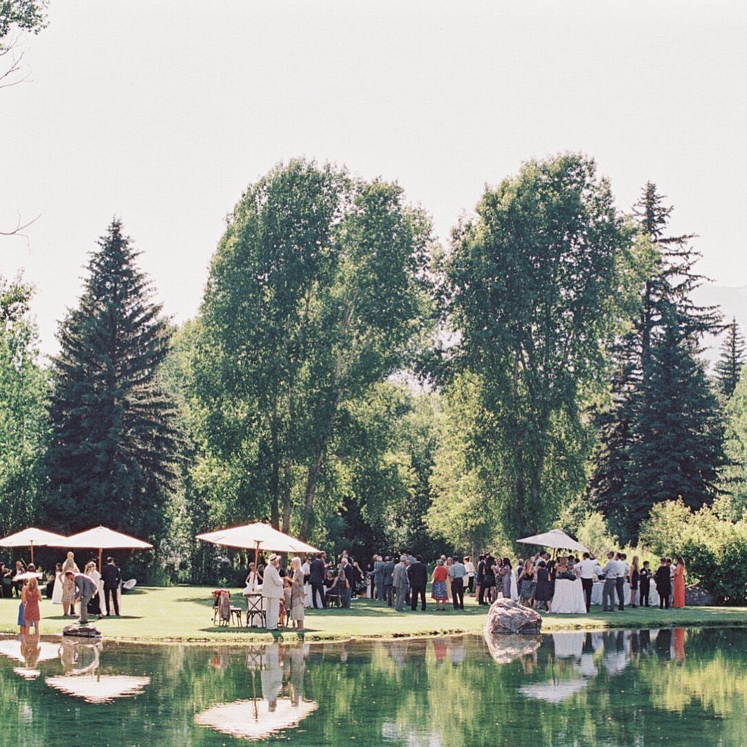 Formal wedding guests gathering on a lawn by a pond with white umbrellas.