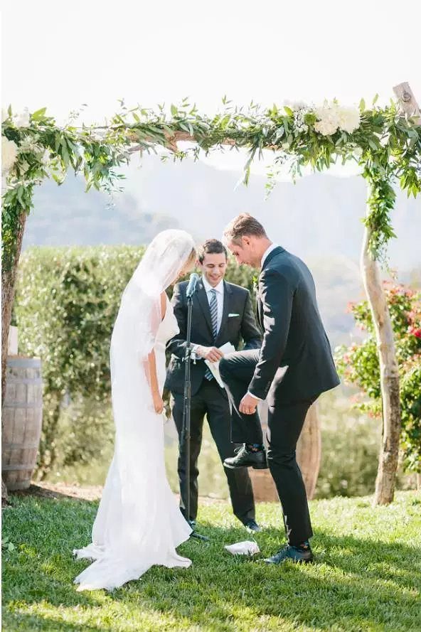 Groom breaking a glass wrapped in cloth under a floral wedding canopy outdoors.