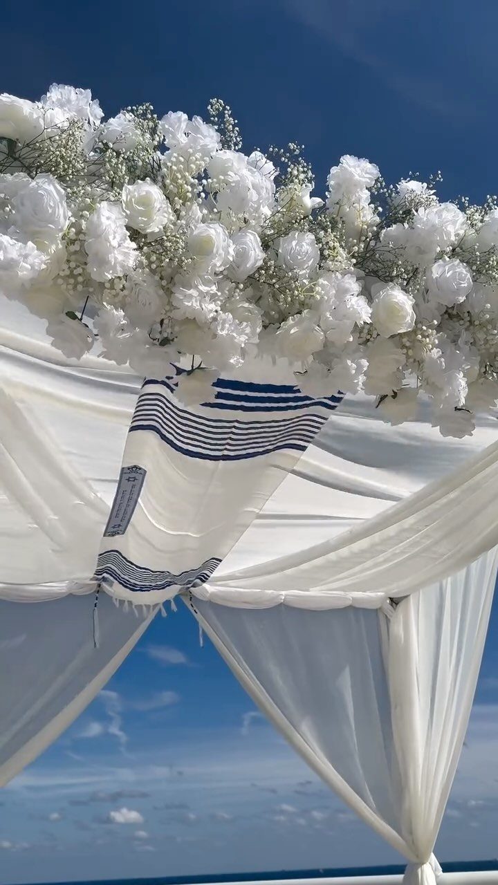 White wedding canopy decorated with white flowers and a blue striped tallit prayer shawl.