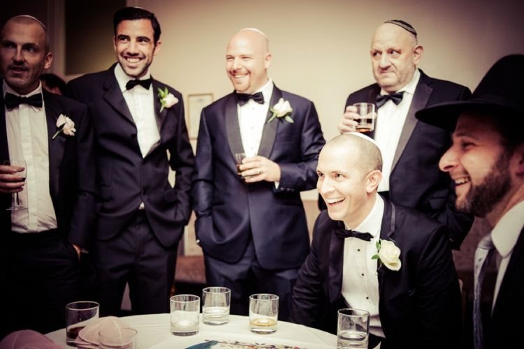 Groom and male guests in tuxedos gathering around a table for a Tisch celebration.