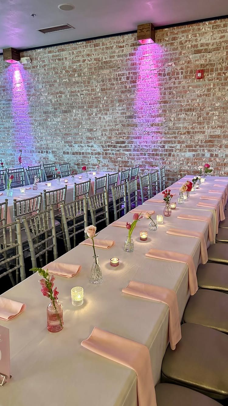 Wedding reception tables with white linens, pink napkins, and purple lighting against brick.