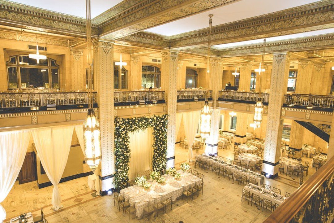 Grand ballroom with gold Art Deco architecture, banquet tables, and a floral wedding arch.