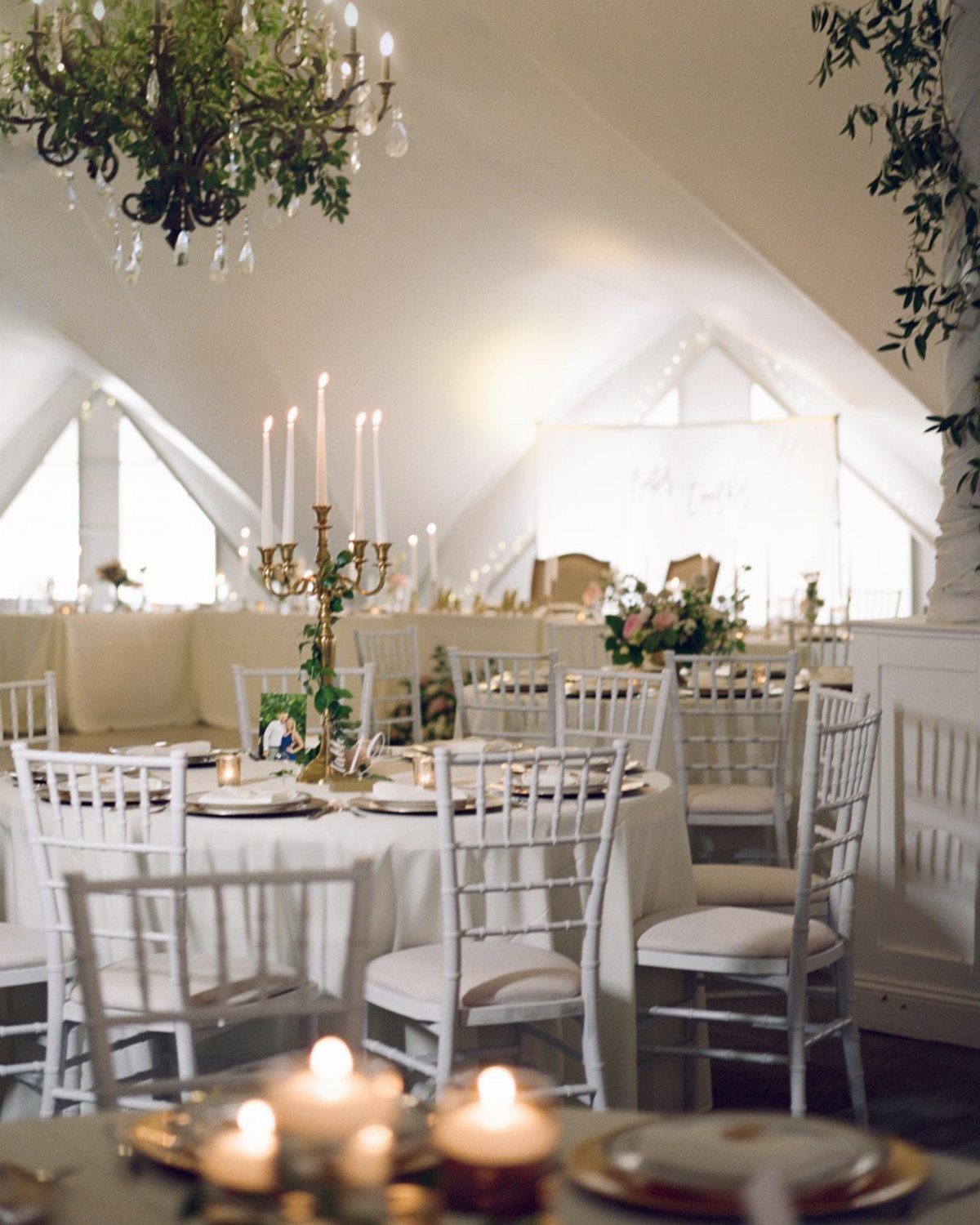 Wedding reception tables with white chairs and gold candelabras inside a white tent.