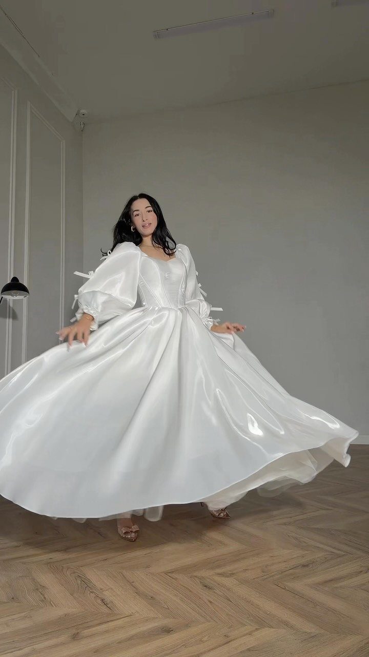 Woman in a voluminous white satin wedding gown posing in a bright room.