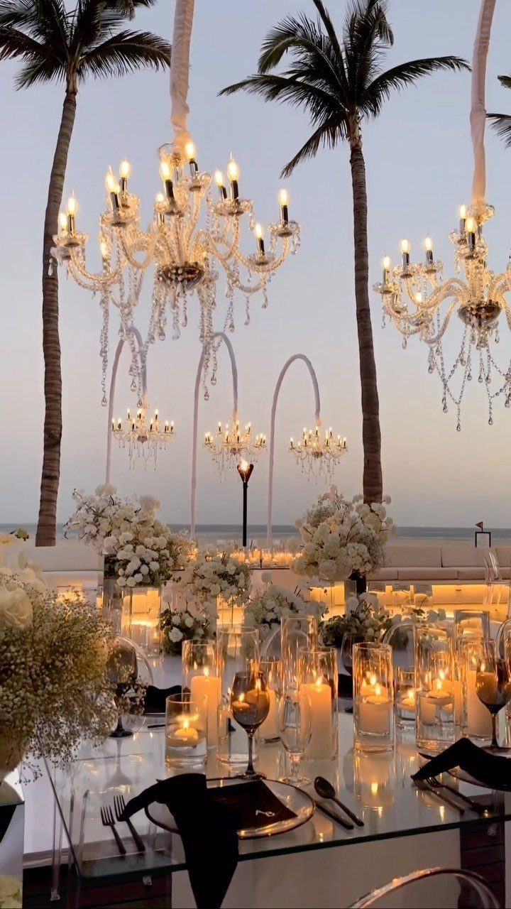 Elegant beach wedding reception table with crystal chandeliers, white flowers, and glowing candles.