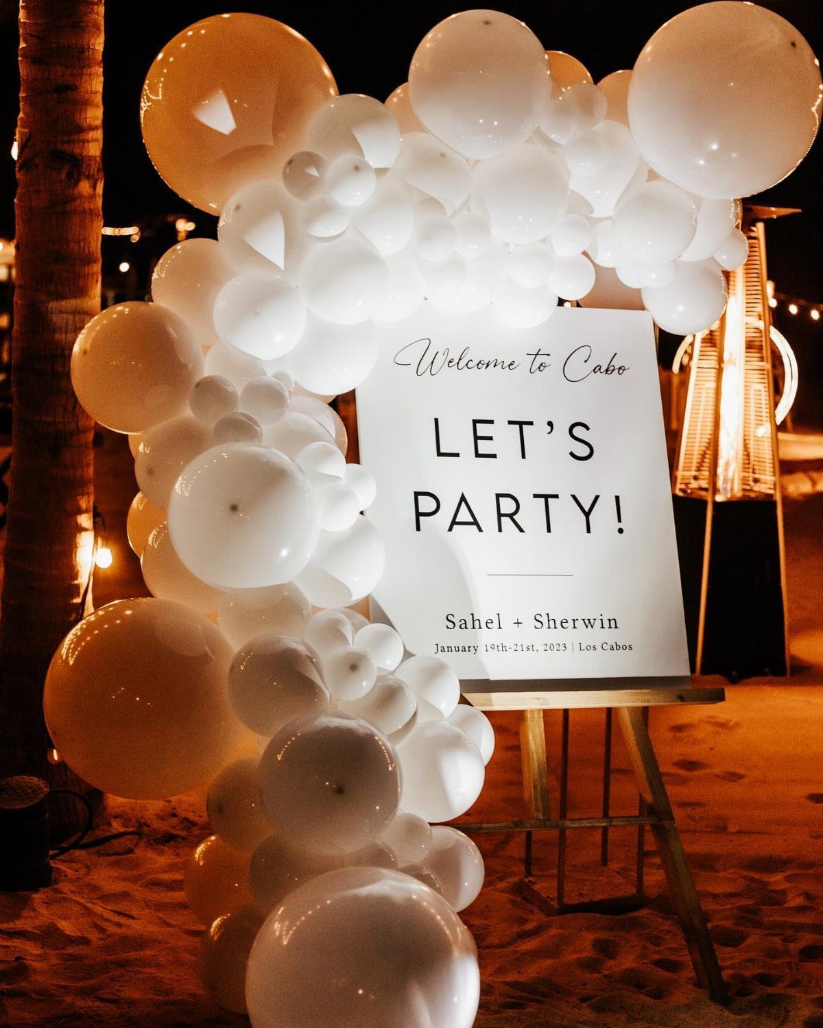 White balloon arch and welcome sign on a sandy beach at night.
