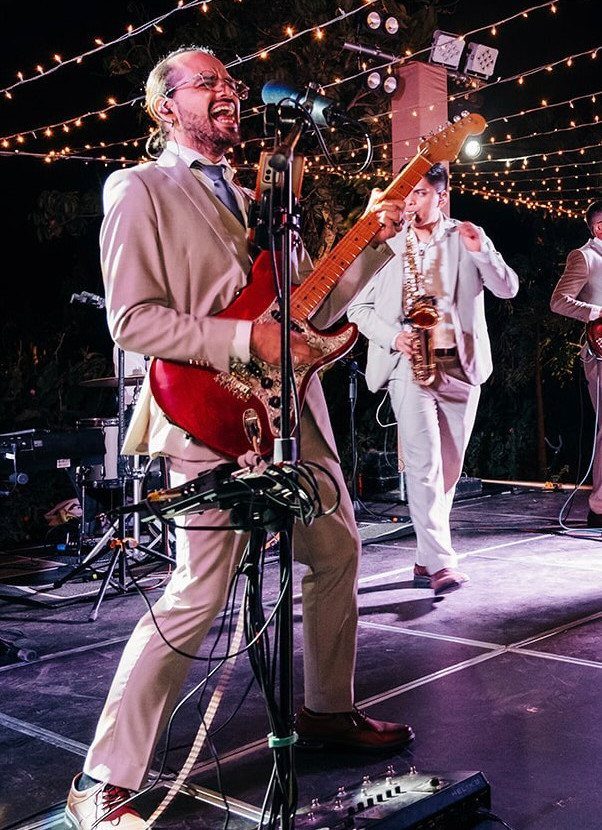 Musicians in suits performing with electric guitar and saxophone on a lit outdoor stage