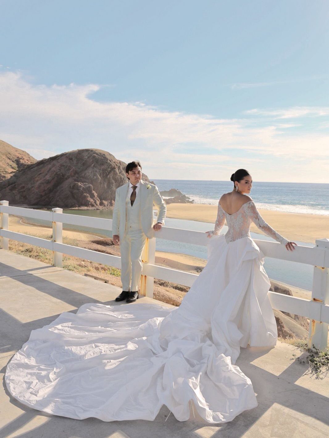 Bride in white lace gown and groom in white suit by beach coastline