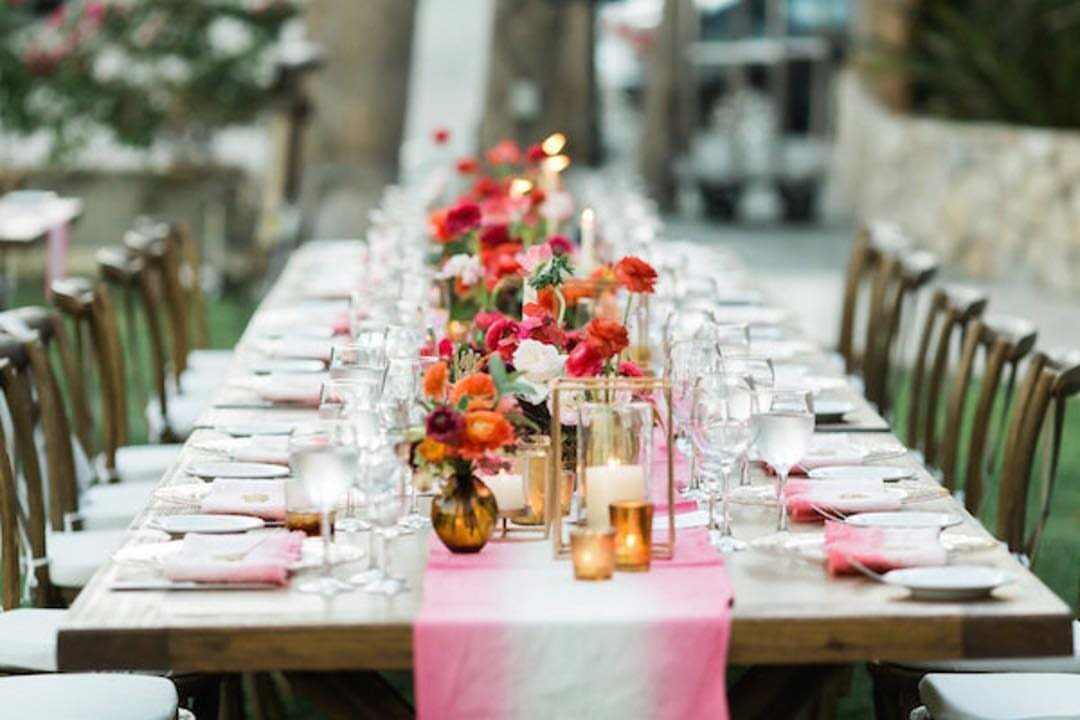Long wooden wedding table with pink runner and vibrant floral centerpieces outdoors