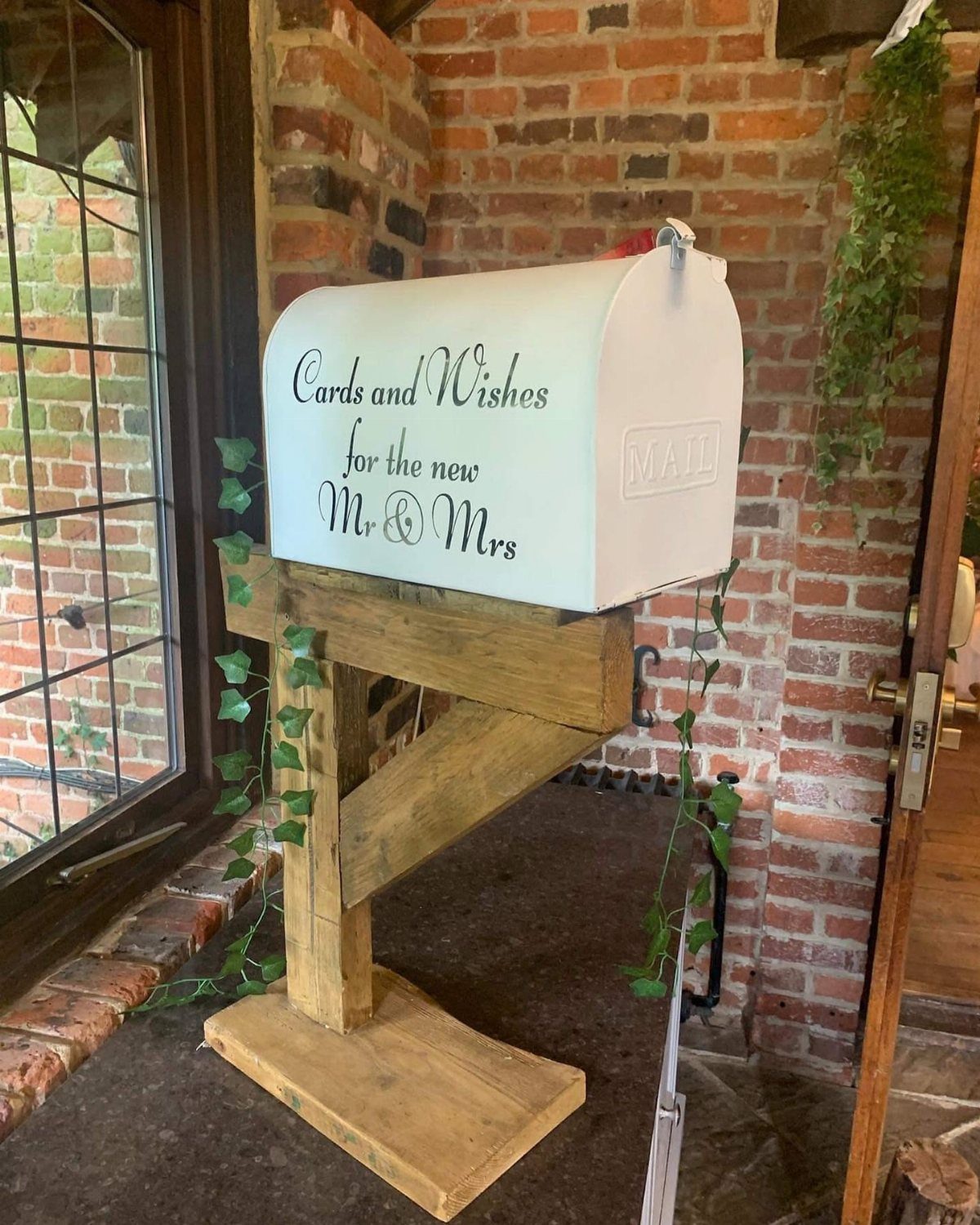 White wedding mailbox on a wooden stand with vines against a brick wall