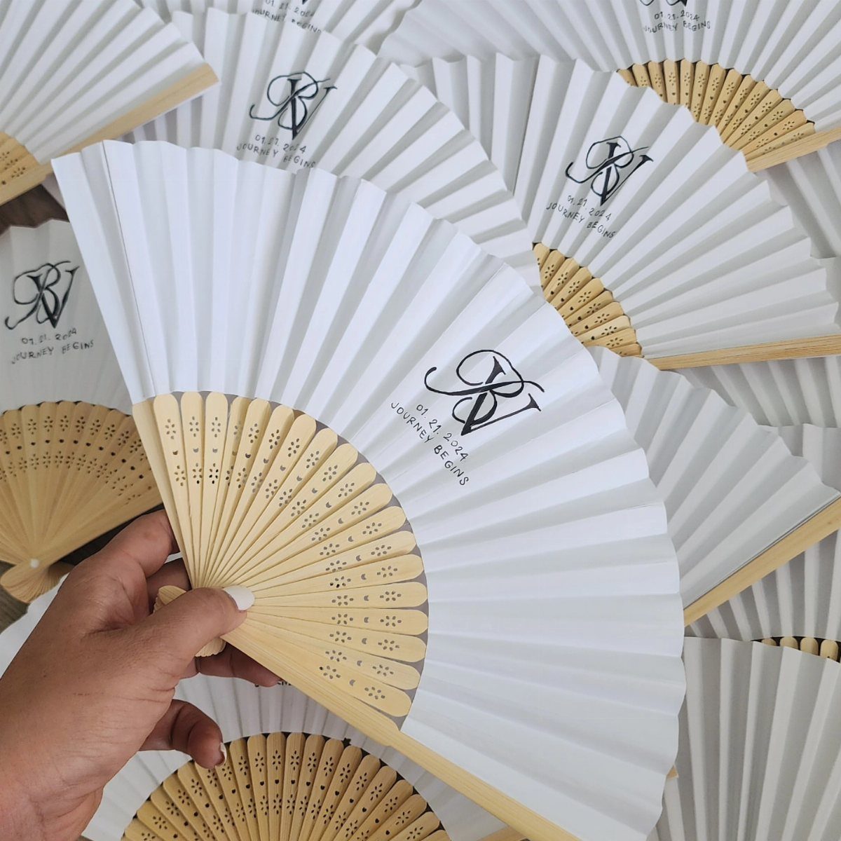 White folding fan with a black monogram and wooden ribs held by hand.