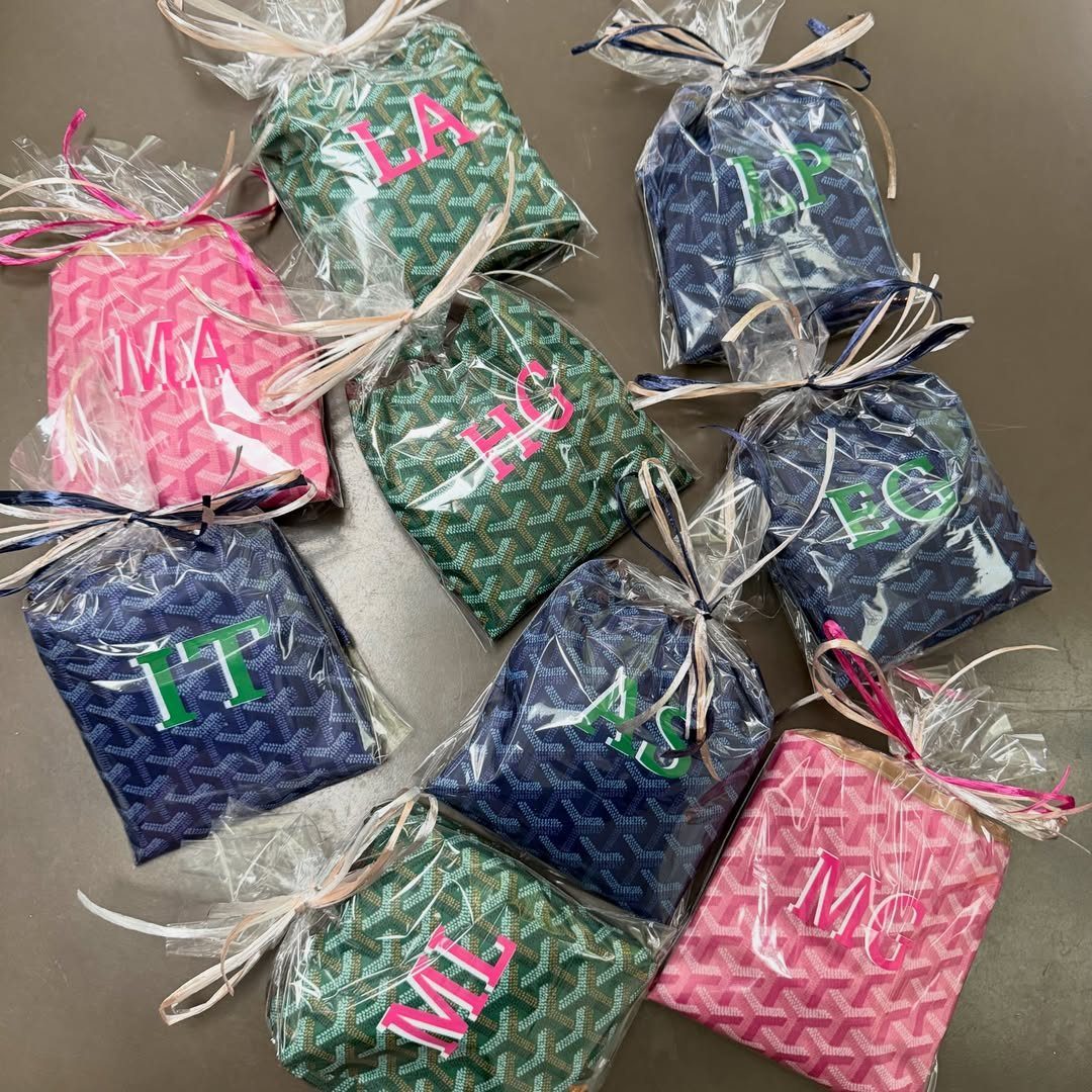 Monogrammed tote bags in pink, green, and navy blue wrapped in clear plastic.