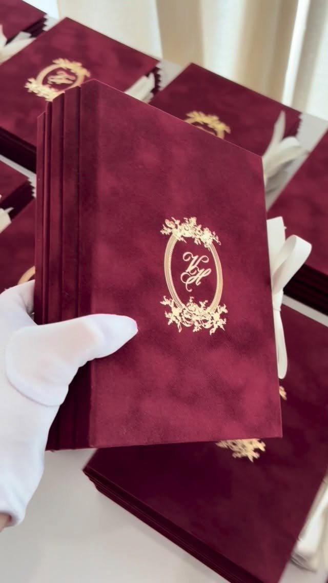 Burgundy velvet wedding invitations with gold foil monograms held by a gloved hand.
