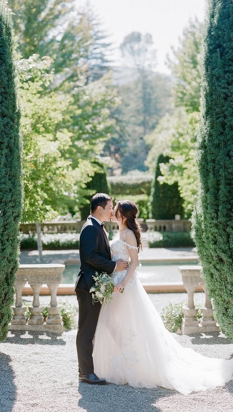 Bride and groom kissing in a formal garden with gravel paths and cypress trees.
