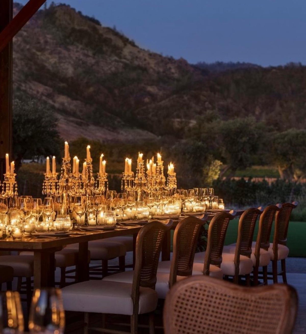 Elegant outdoor wedding dinner table with glowing candles and cane chairs against hills.