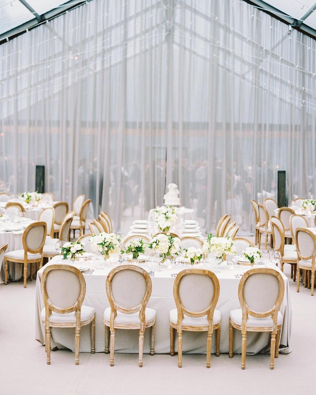 Wedding reception tables with white flowers and cream chairs inside a sheer tent