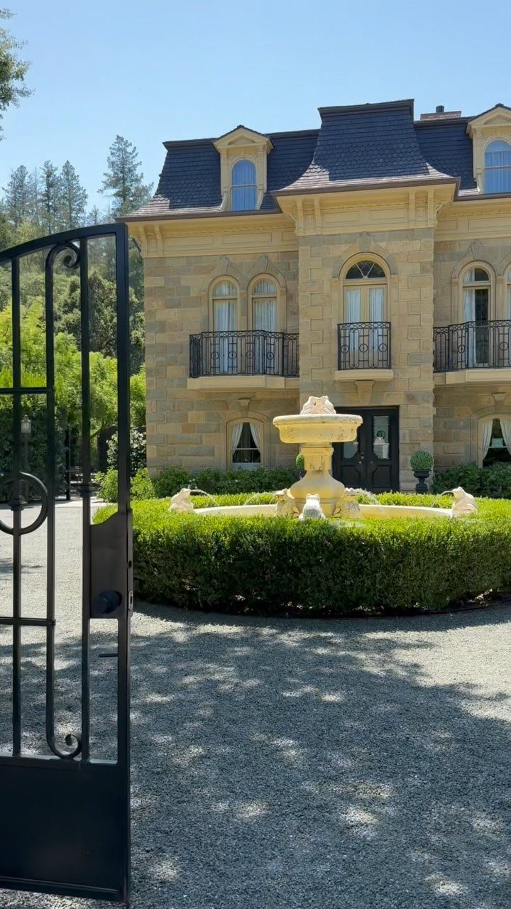 Stone manor house with a tiered fountain and manicured hedge behind iron gates.