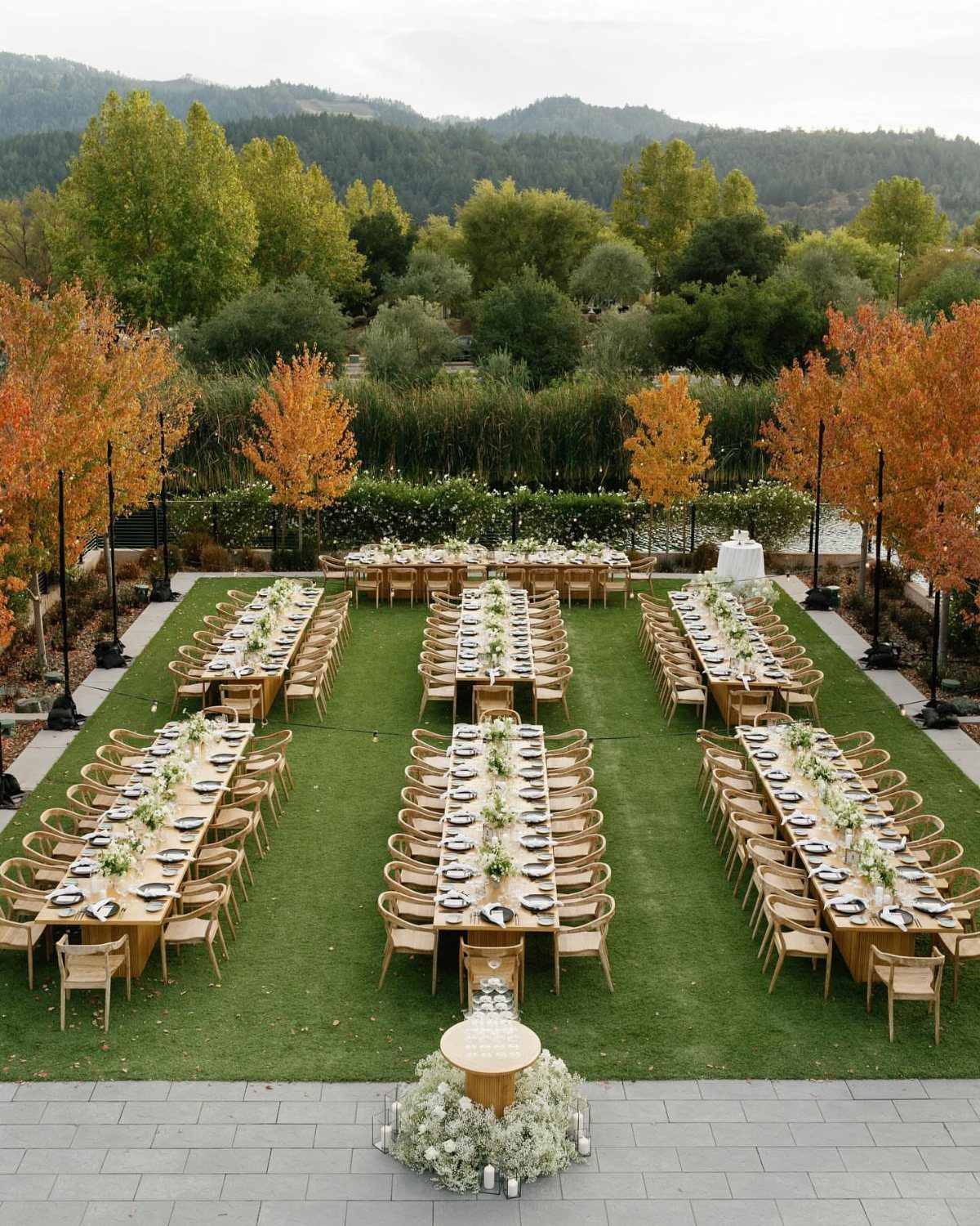 Long wooden wedding tables set on a green lawn with autumn trees and mountains.