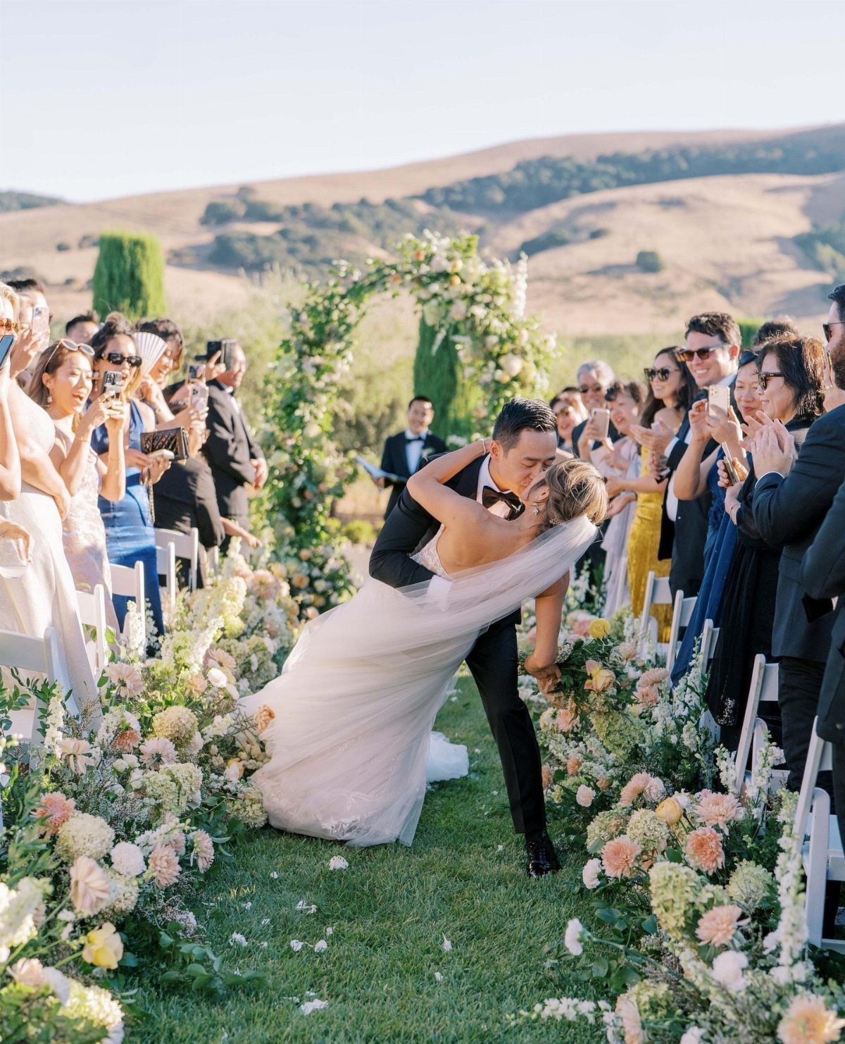 Groom dipping and kissing bride during an outdoor vineyard wedding ceremony with guests.