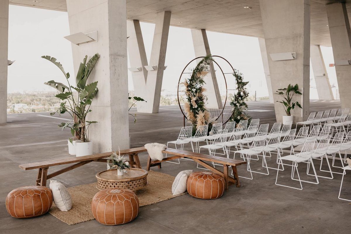 Modern wedding ceremony setup with white chairs, leather poufs, and a circular floral arch.