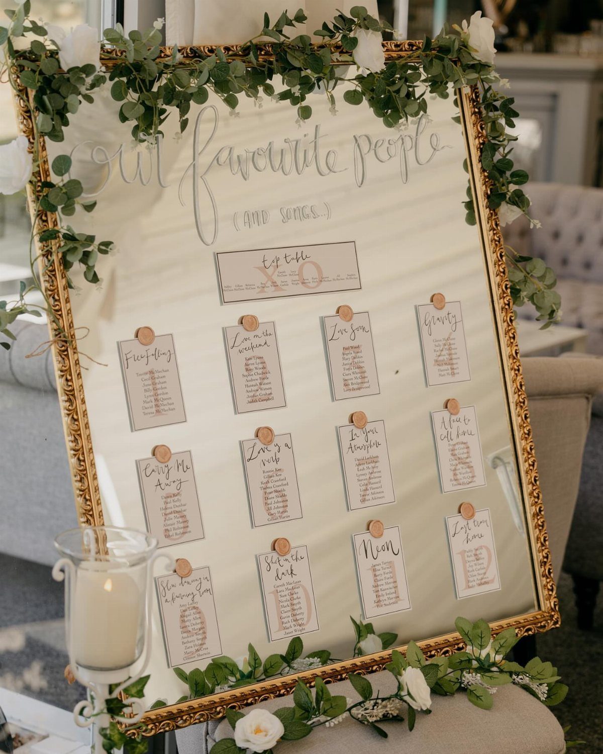 Gold-framed wedding seating chart decorated with green leaves and white floral accents.