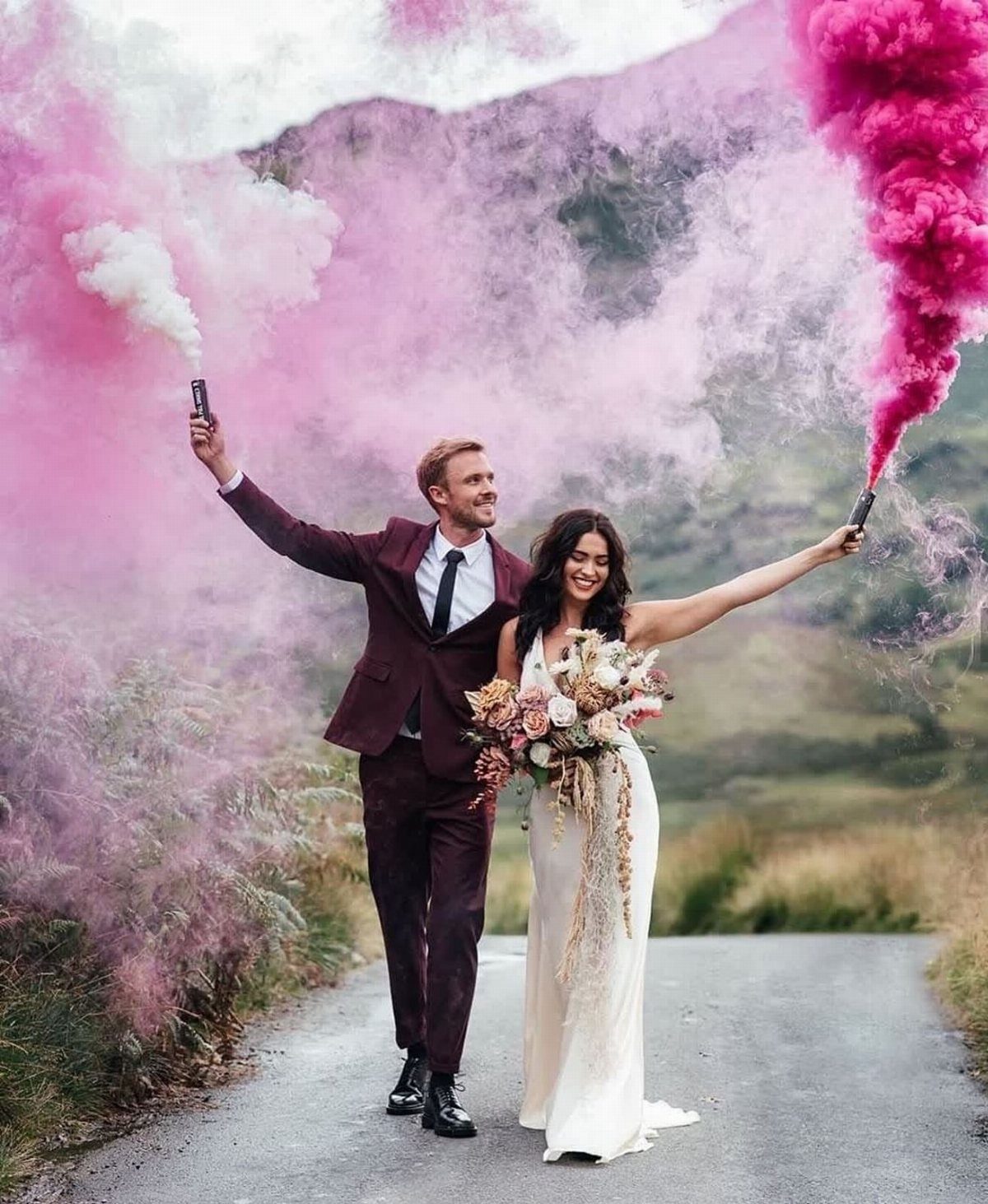 Bride and groom holding smoke bombs emitting pink and white smoke on a road.