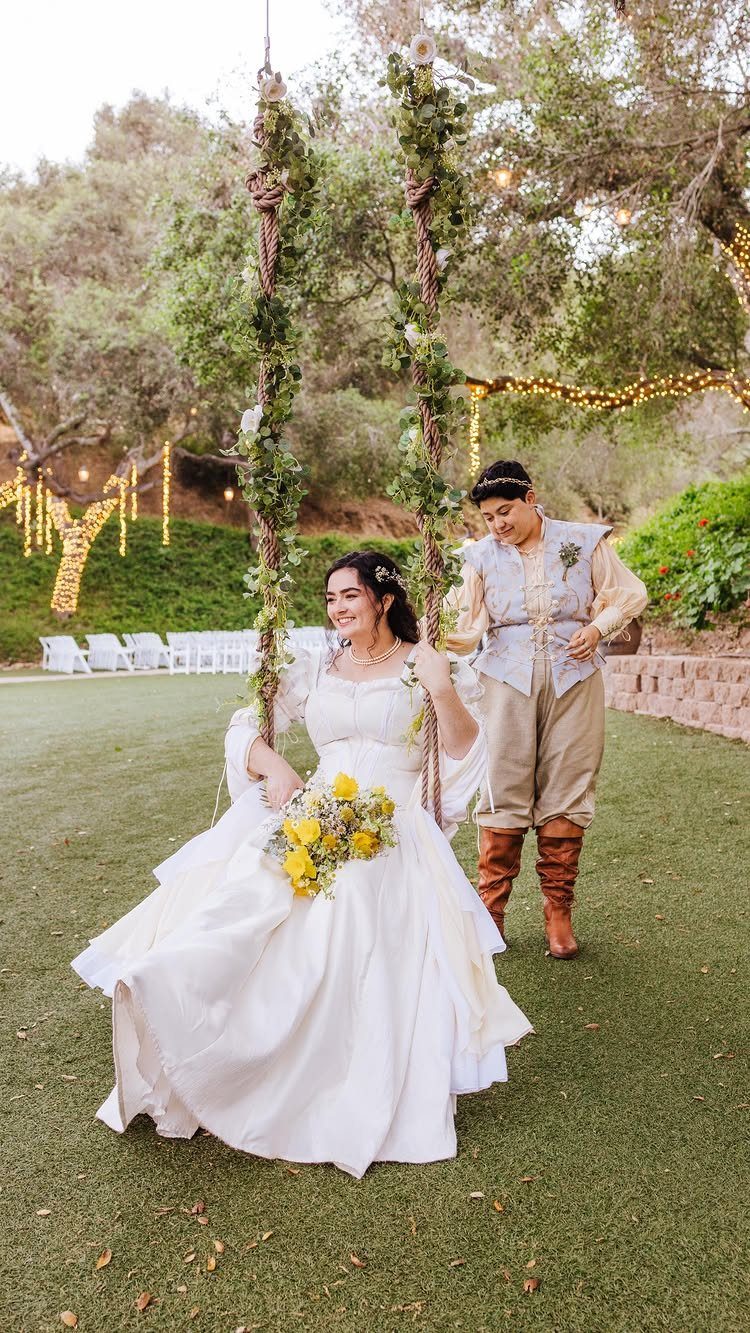 Bride and groom in medieval themed costumes posing on a floral swing outdoors.