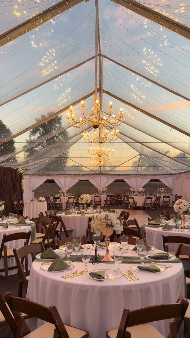 Wedding reception tables with white linens and gold chandeliers inside a clear tent.