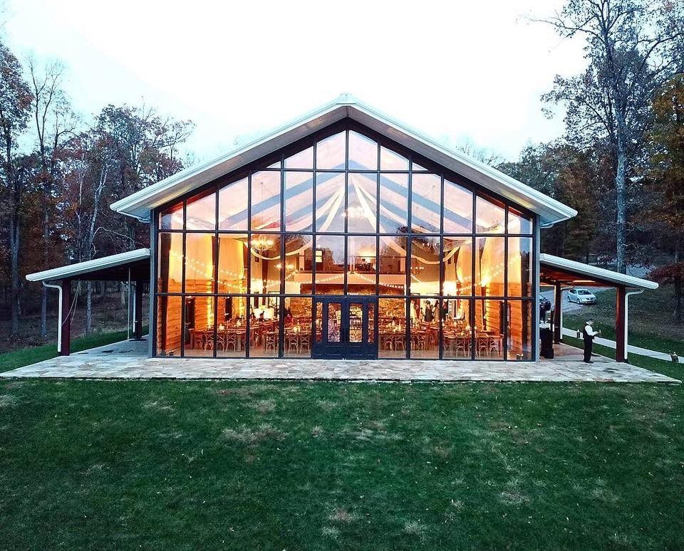Glass-walled event pavilion glowing with warm light at dusk on a green lawn