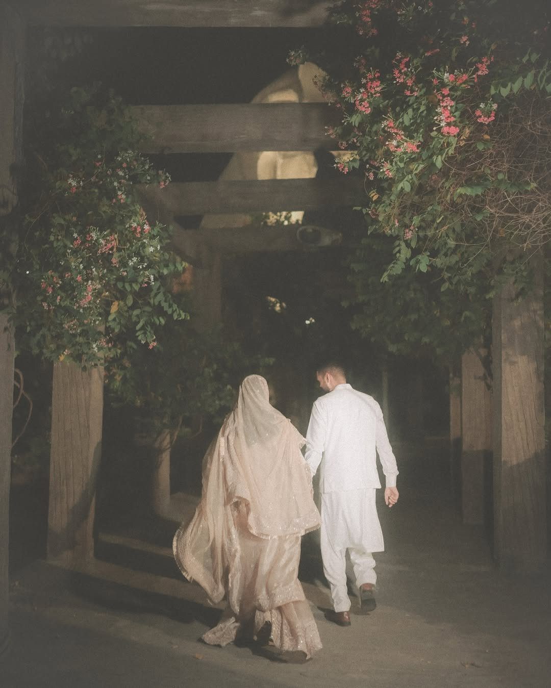 Couple in traditional wedding attire walking through a wooden pergola at night.