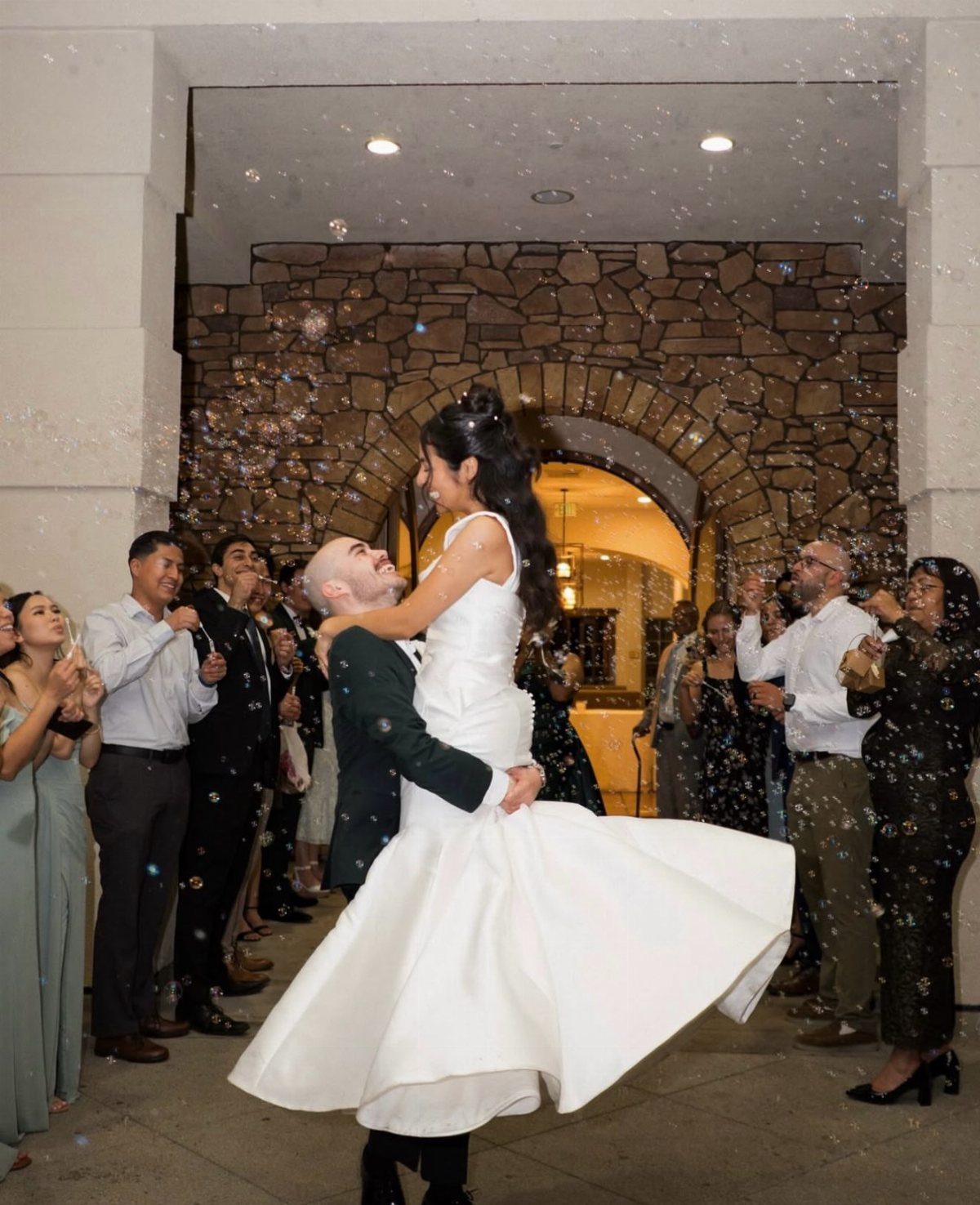Groom lifting bride in white gown surrounded by guests and bubbles at night.
