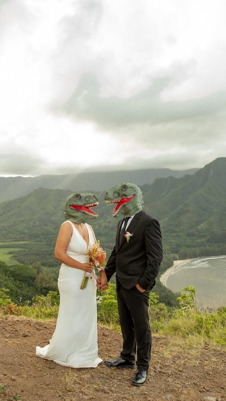 Couple in dinosaur masks wearing wedding attire standing on a scenic tropical cliffside.
