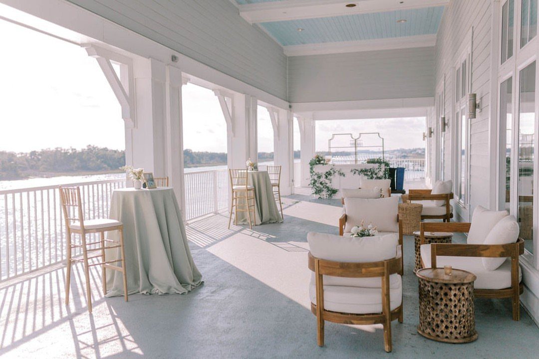 White covered porch with lounge seating and draped tables overlooking a coastal waterfront.