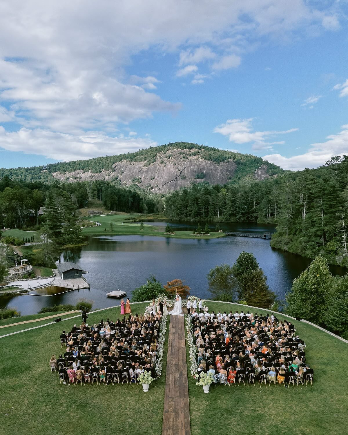 Outdoor wedding ceremony with guests seated on a lawn overlooking a lake and mountains.