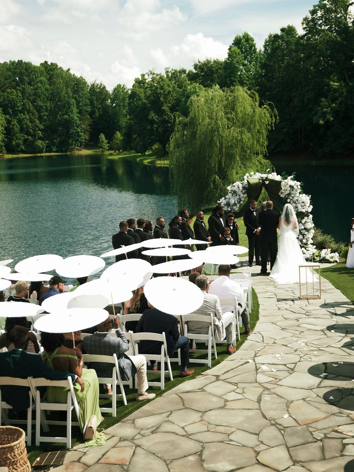 Wedding ceremony by a lake with guests holding white parasols under green trees