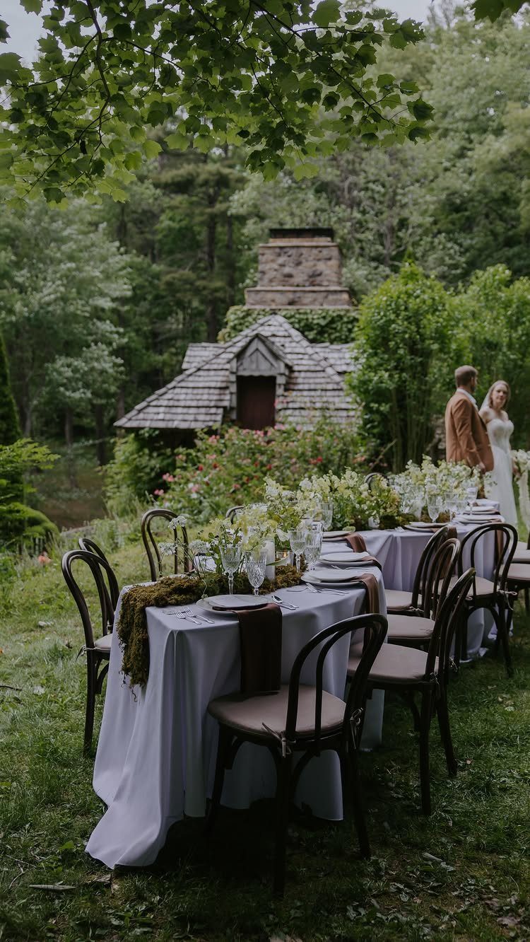 Outdoor wedding reception table with white linens and a couple by a rustic cottage