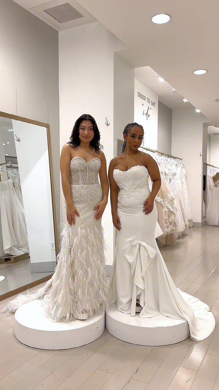 Two women wearing glamorous white strapless wedding gowns in a bright bridal boutique.
