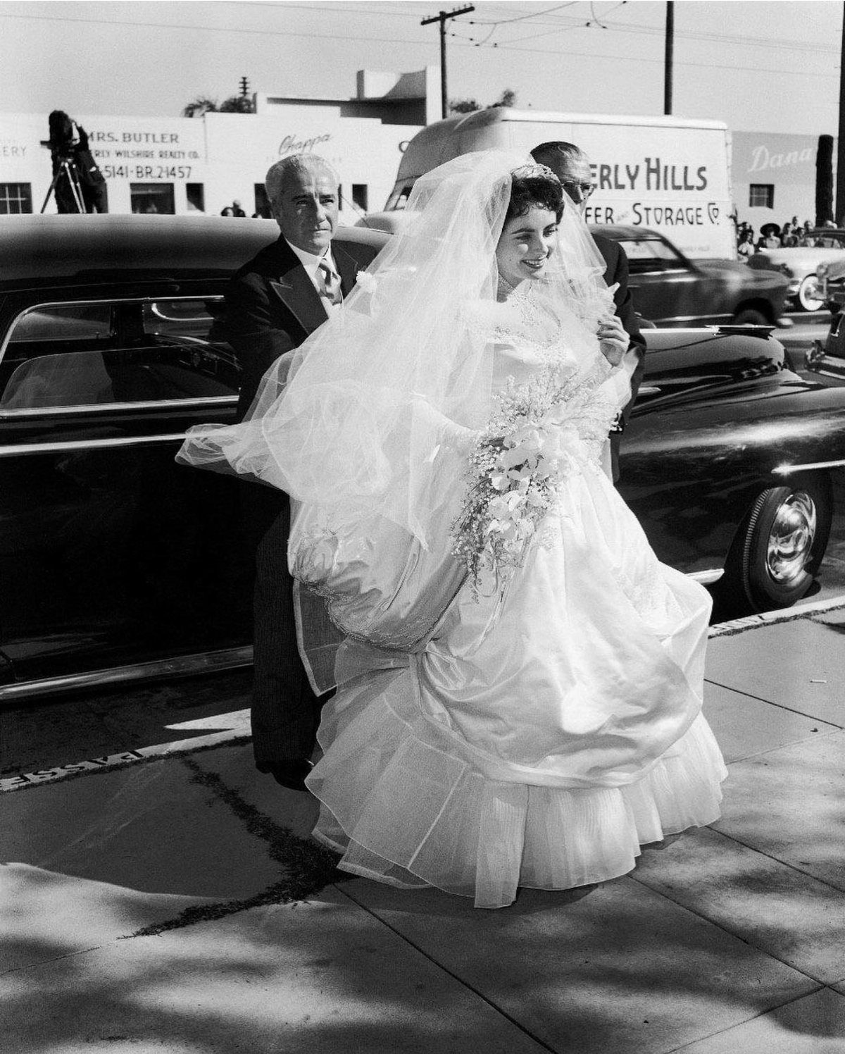 Bride in a voluminous white gown and veil stepping out of a vintage car.