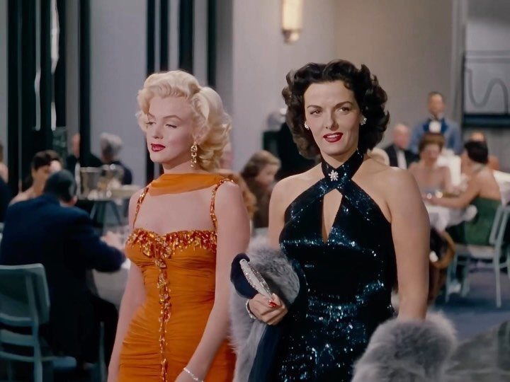 Two glamorous women in vintage evening gowns and retro hairstyles in a dining room.