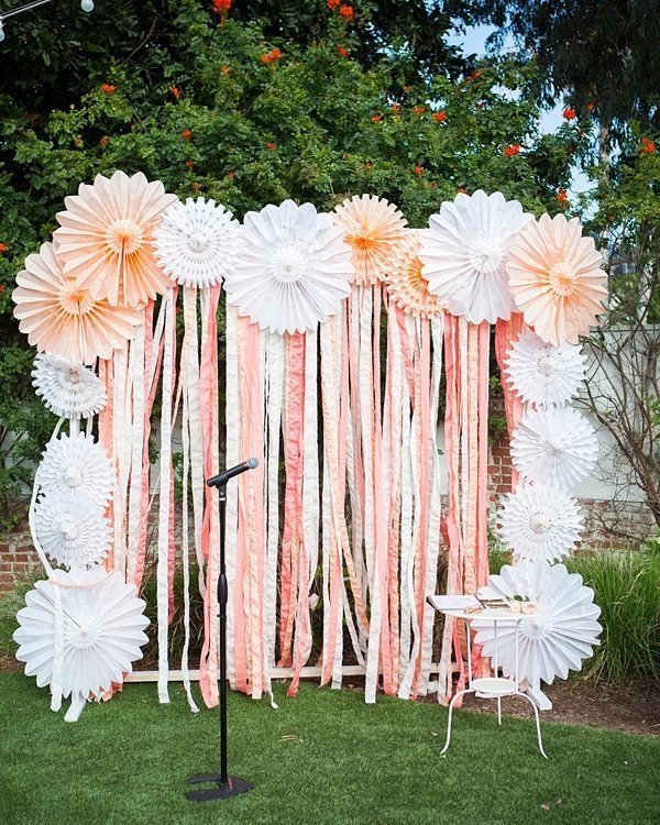 Peach and white paper fan backdrop with ribbons and a microphone stand outdoors.