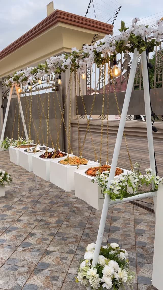 Food platters on white pedestals under a floral canopy with hanging lights outdoors.