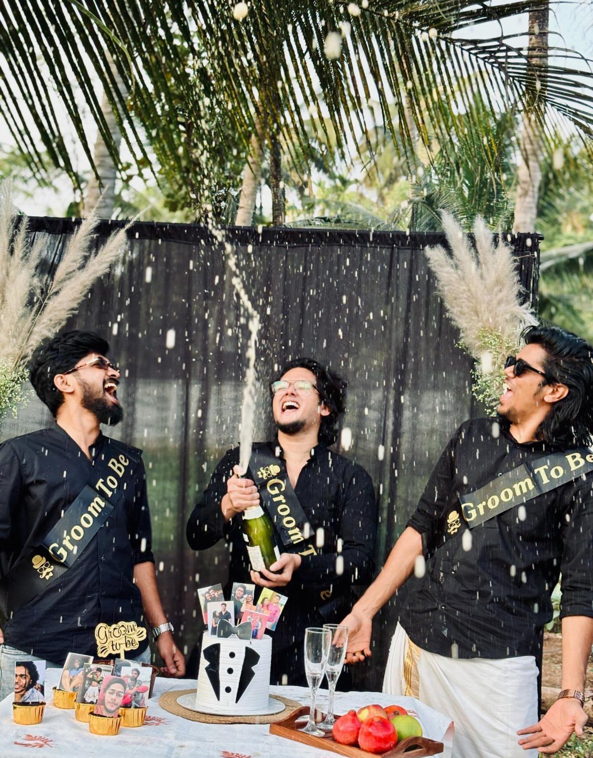 Three men celebrating a bachelor party with champagne, a tuxedo cake, and sashes.