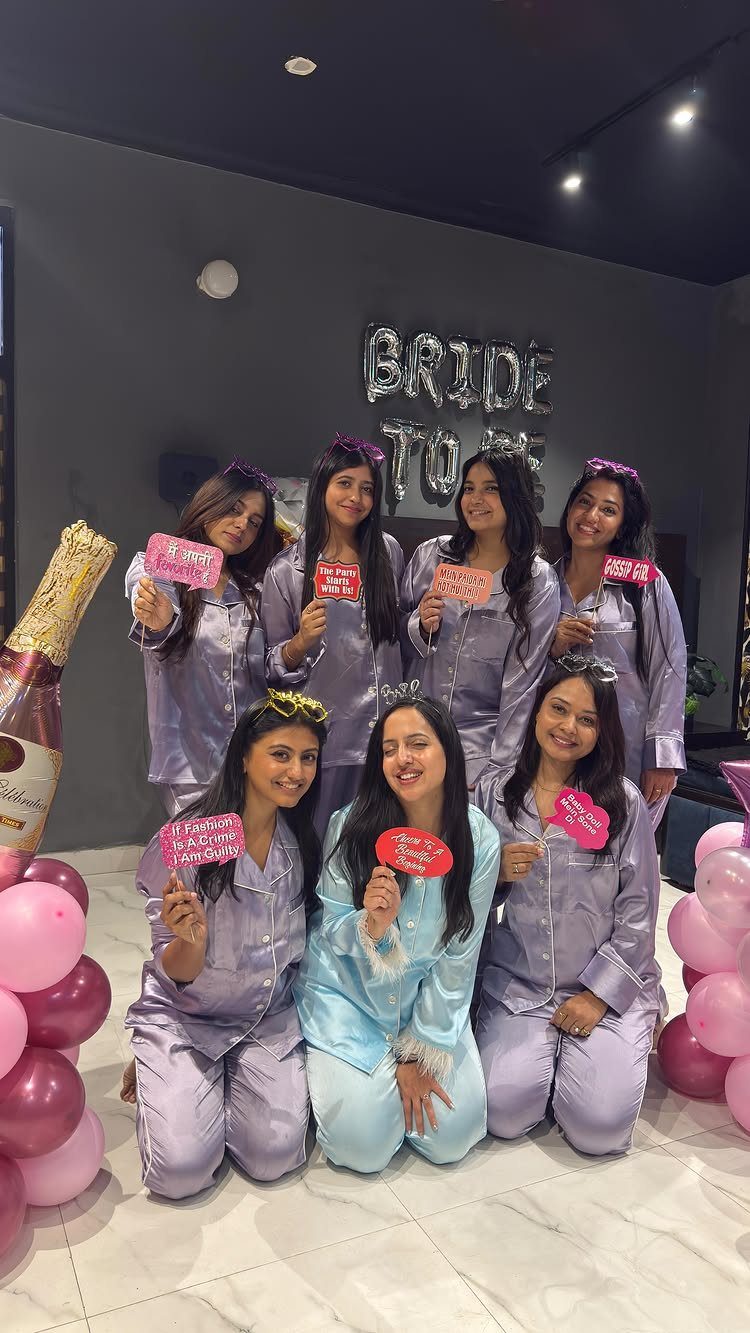 Seven women in satin pajamas posing with 'BRIDE TO BE' balloons and pink hearts.