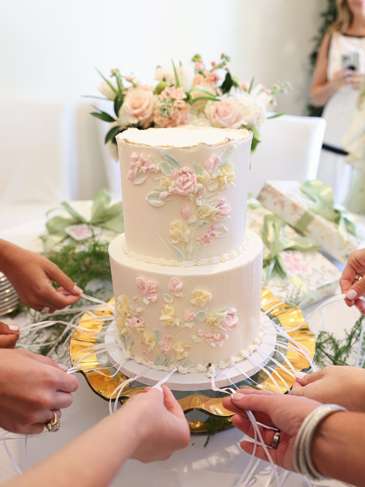 Two-tier white floral cake with ribbons being held by guests at a party