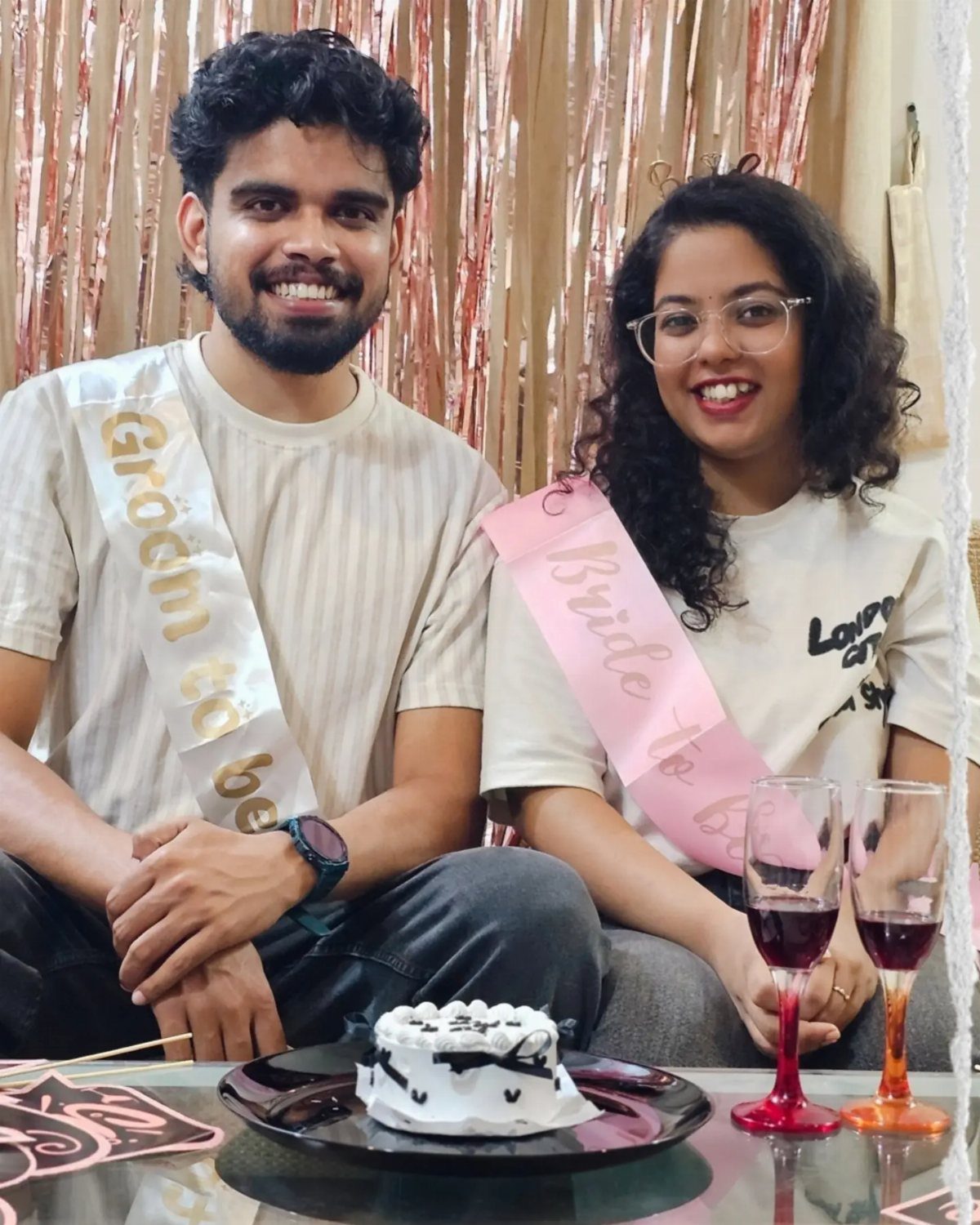 Couple wearing bride and groom sashes sitting with a cake and wine.