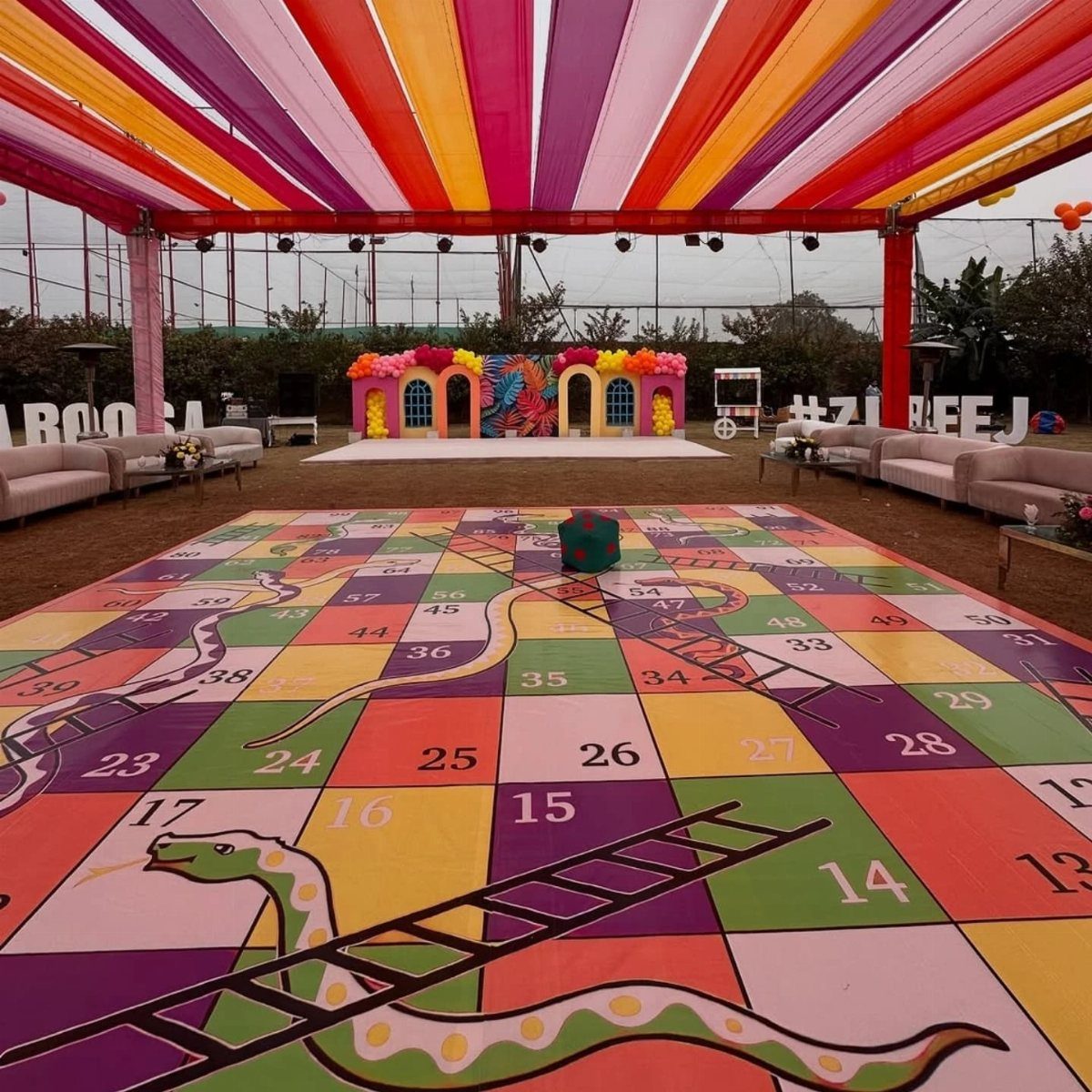 Giant snakes and ladders game board on grass under a colorful fabric canopy.