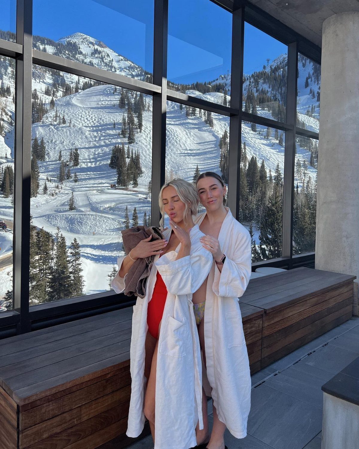 Two women in white bathrobes posing before a window overlooking snowy mountains.